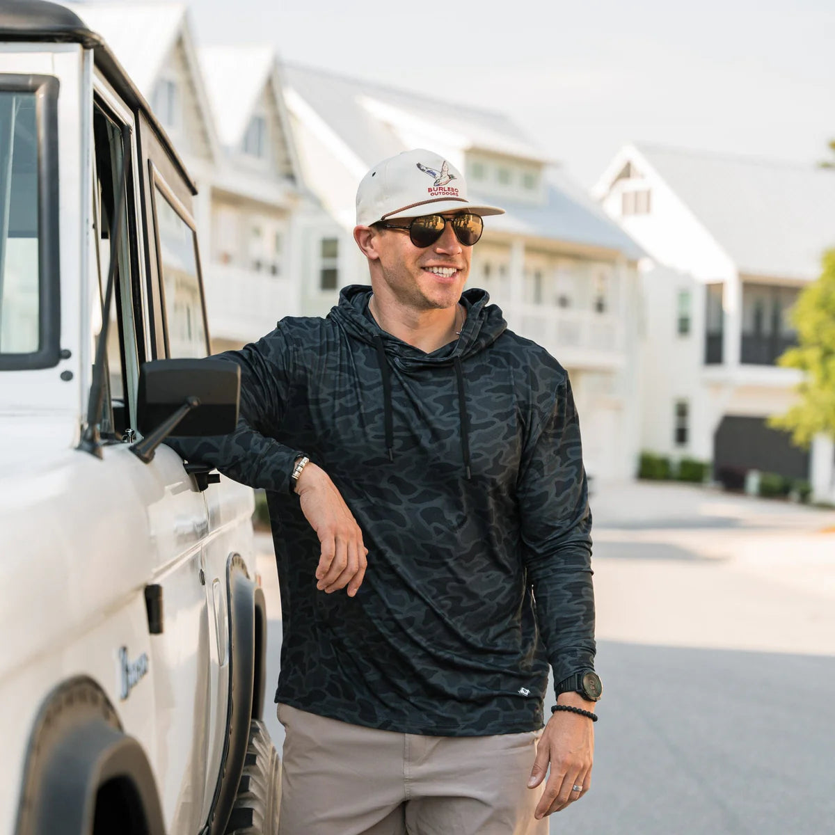 Man leaning against a white vehicle wearing a dark patterned hoodie and beige cap.