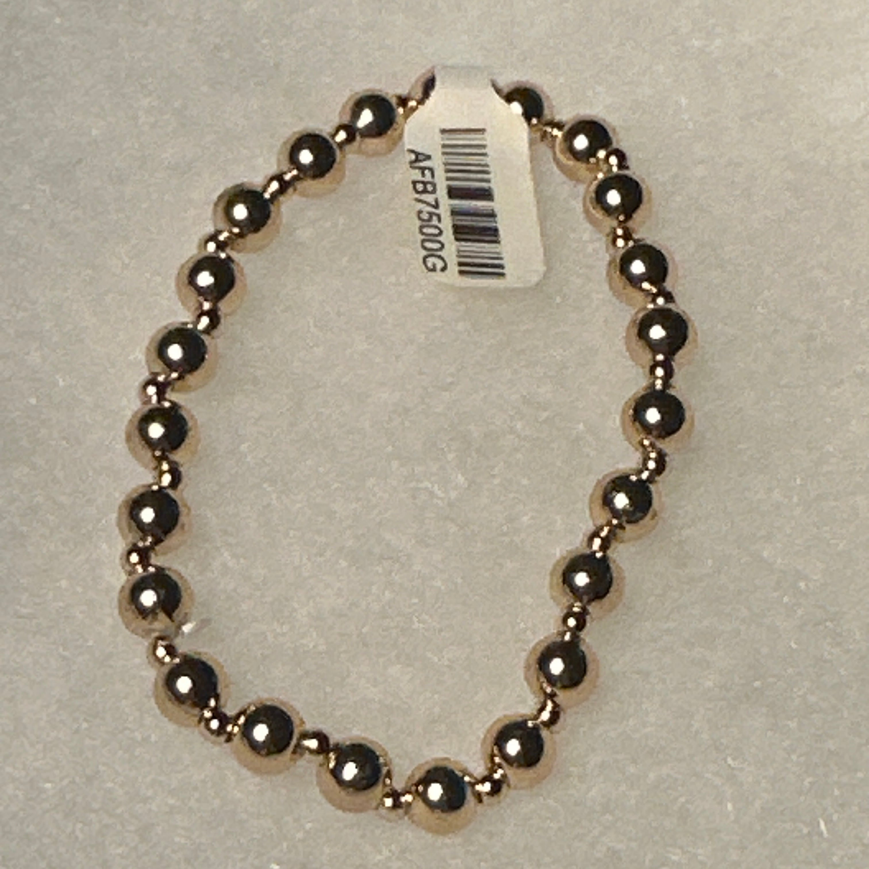 A beaded stretch bracelet made of polished metallic dark gold or rose-gold-toned beads, shown on a light gray textured background with a white product tag attached.