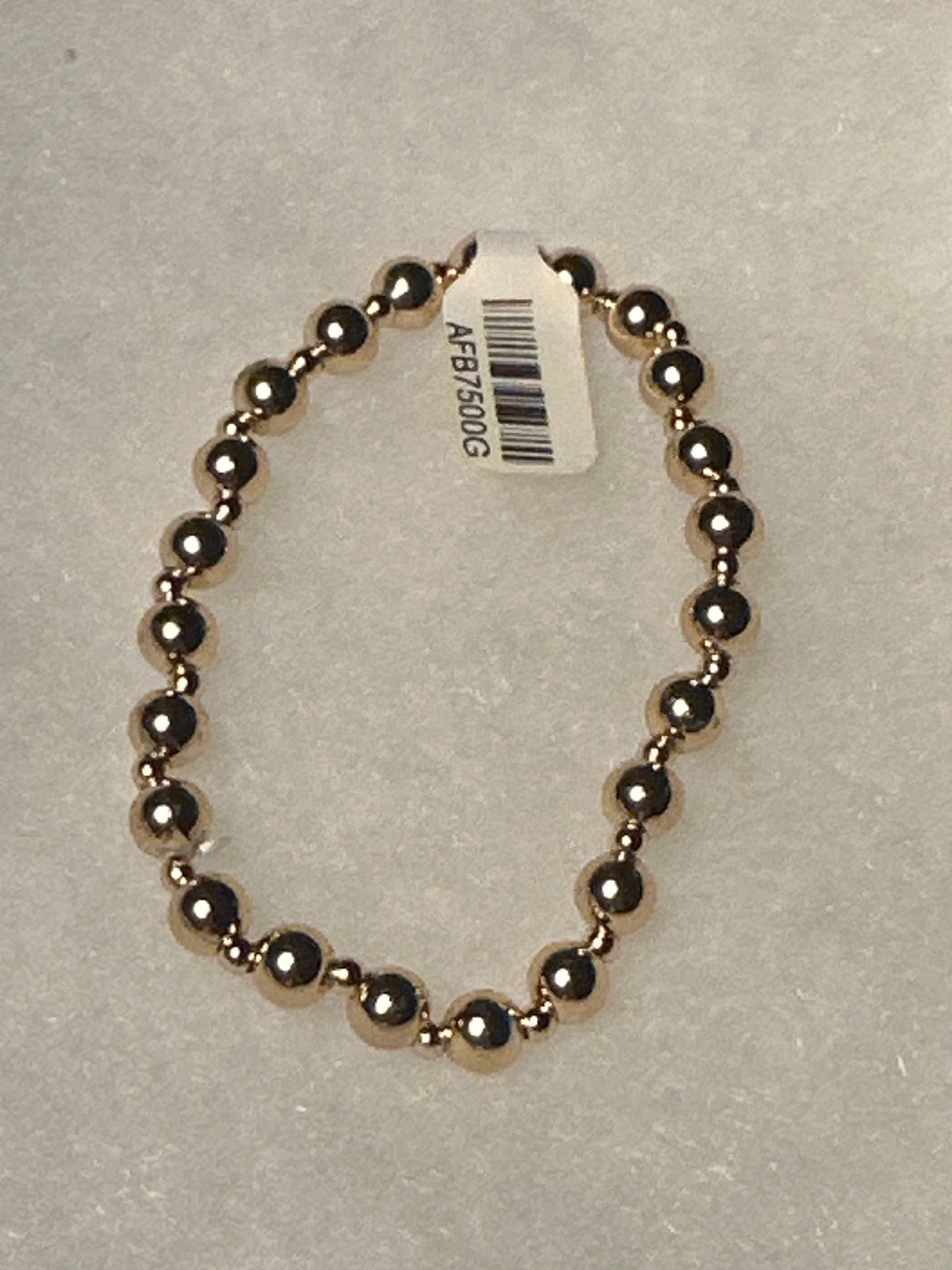 A beaded stretch bracelet made of polished metallic dark gold or rose-gold-toned beads, shown on a light gray textured background with a white product tag attached.