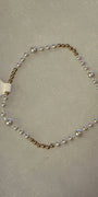 A delicate beaded bracelet featuring alternating white pearls and small gold-toned beads, laid out on a textured light gray surface with a white barcode tag attached.