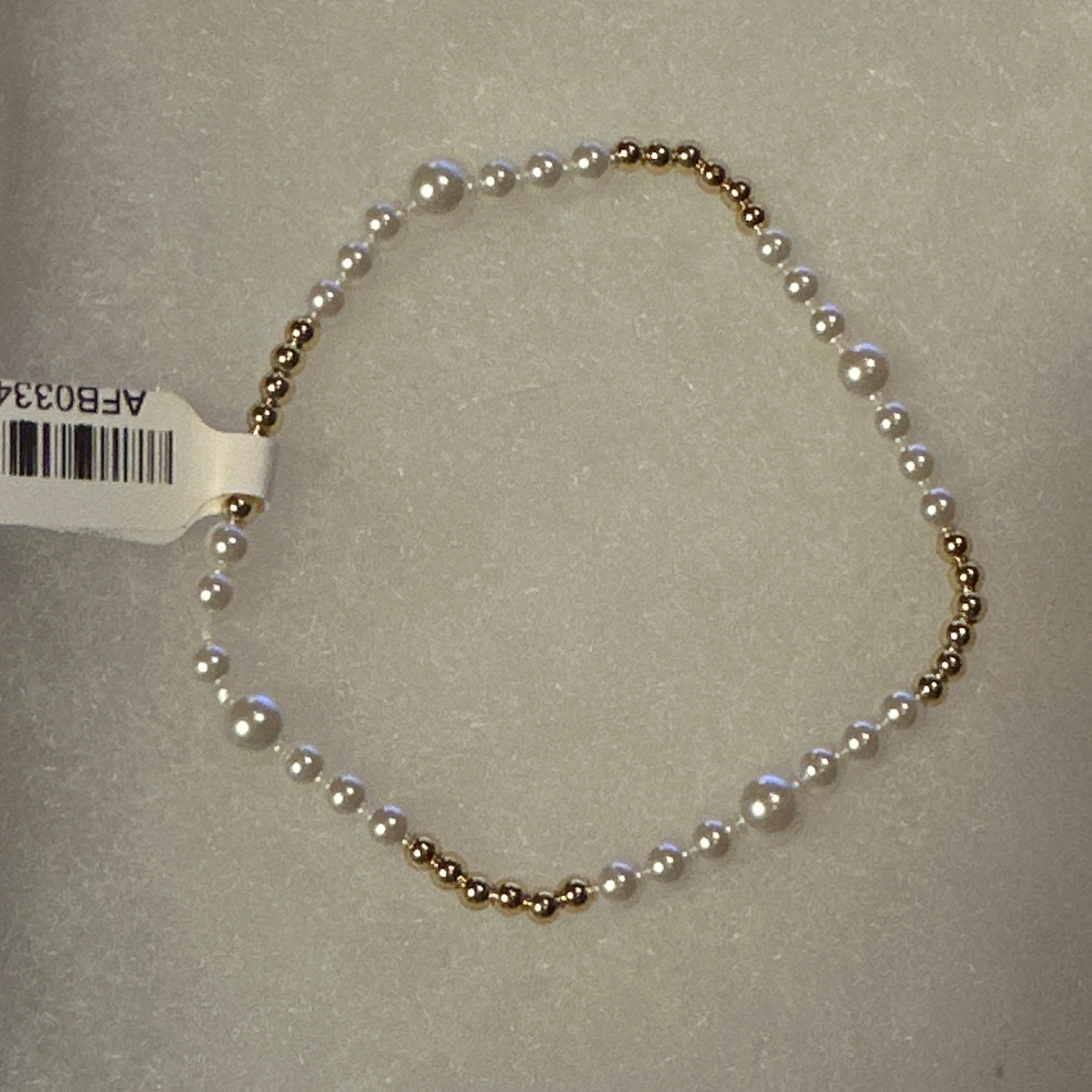 A delicate beaded bracelet featuring alternating white pearls and small gold-toned beads, laid out on a textured light gray surface with a white barcode tag attached.