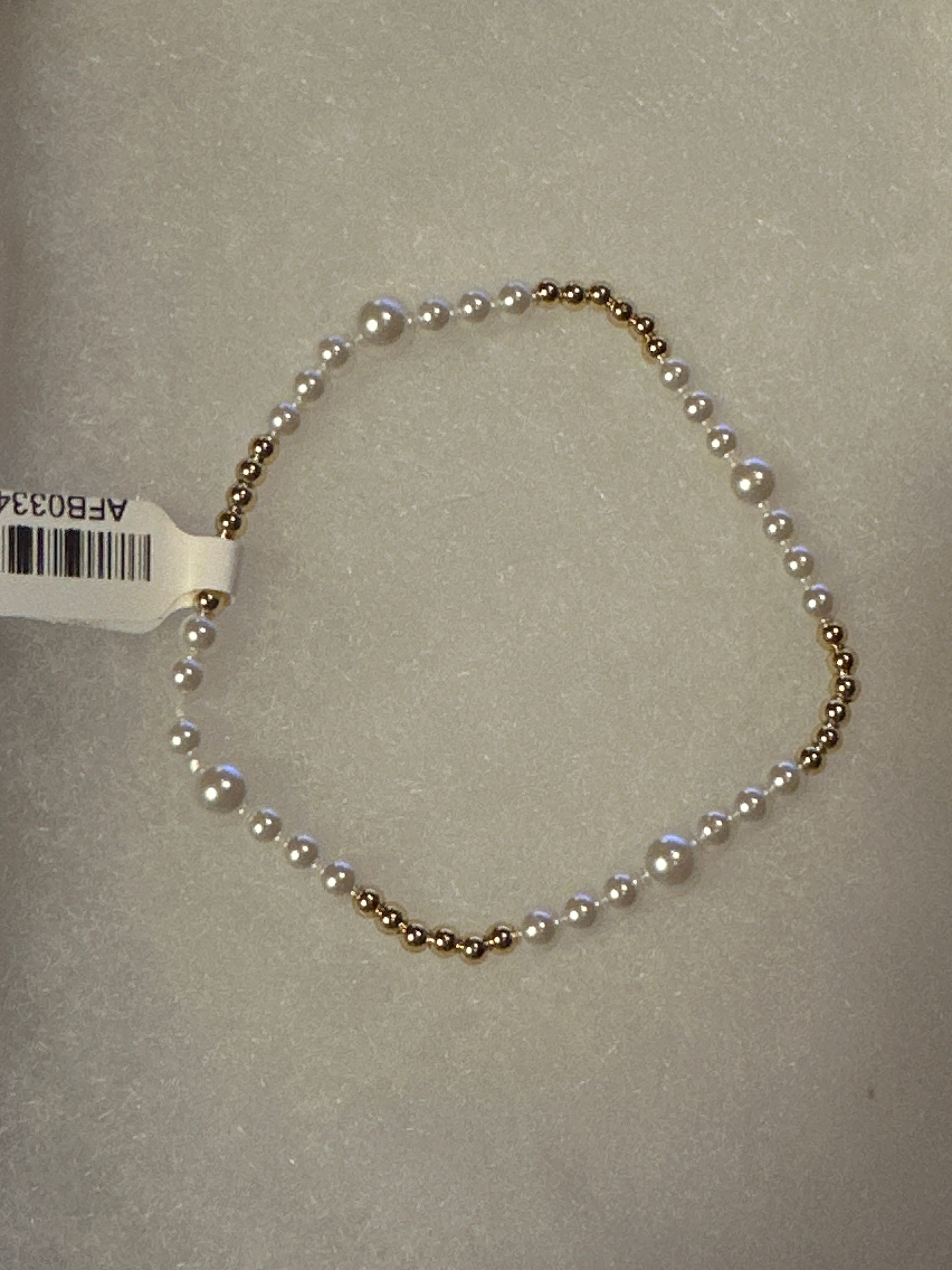 A delicate beaded bracelet featuring alternating white pearls and small gold-toned beads, laid out on a textured light gray surface with a white barcode tag attached.