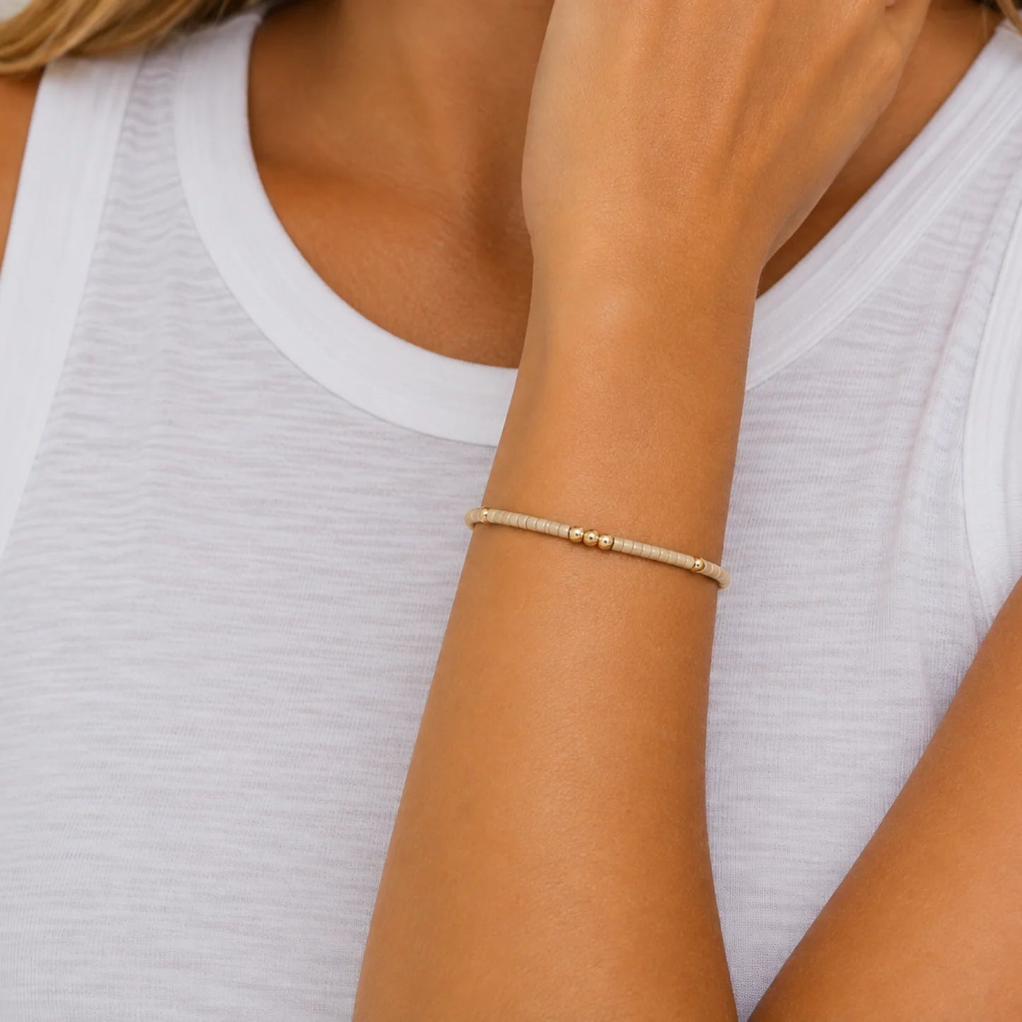 A close-up of a delicate gold beaded bracelet worn on a woman's wrist, paired with a white ribbed tank top.