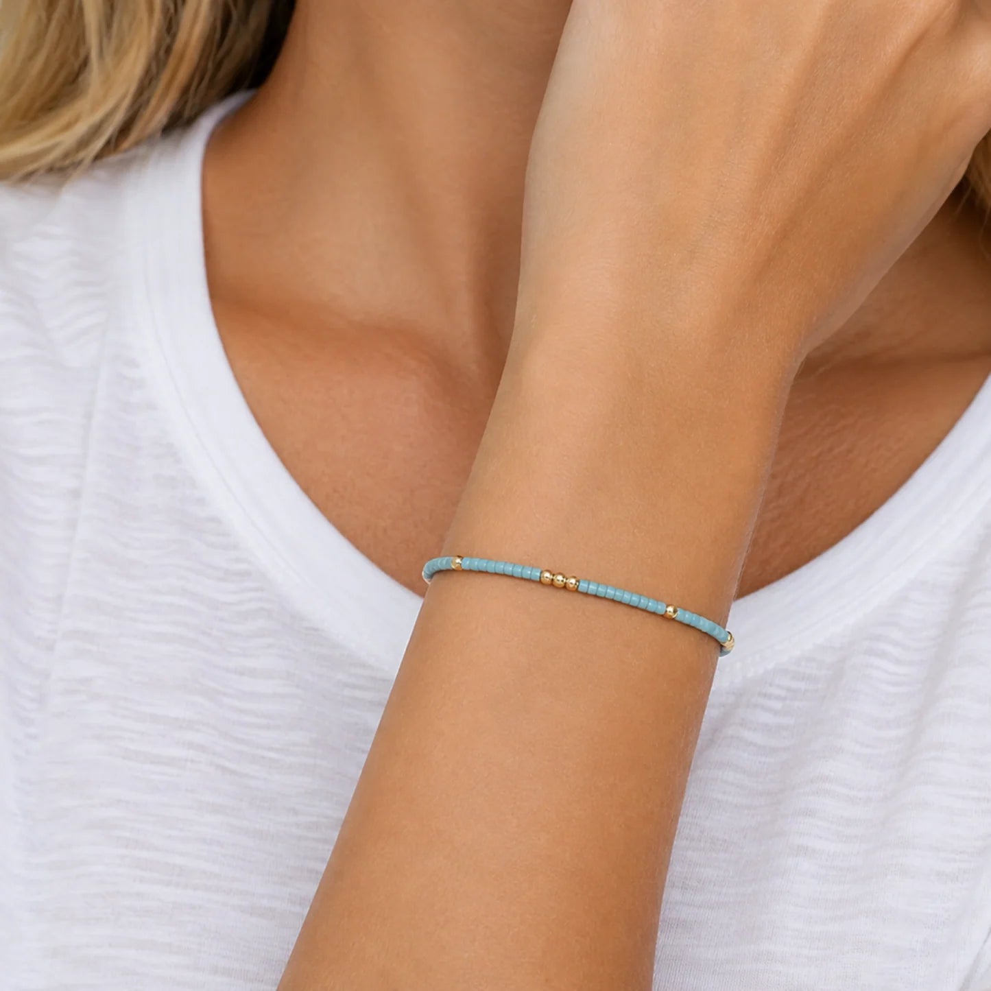 A delicate beaded bracelet with light blue beads and small gold spacer beads worn on a woman's wrist, shown against a white top.