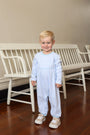 A young toddler with blonde hair stands in front of a white wooden bench wearing a light blue and white checkered long-sleeve footless romper and beige sneakers.