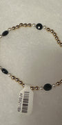 A beaded stretch bracelet with metallic gold-toned round beads, small white pearl-like beads, and faceted black oval beads, shown with a white barcode tag on a light textured background.
