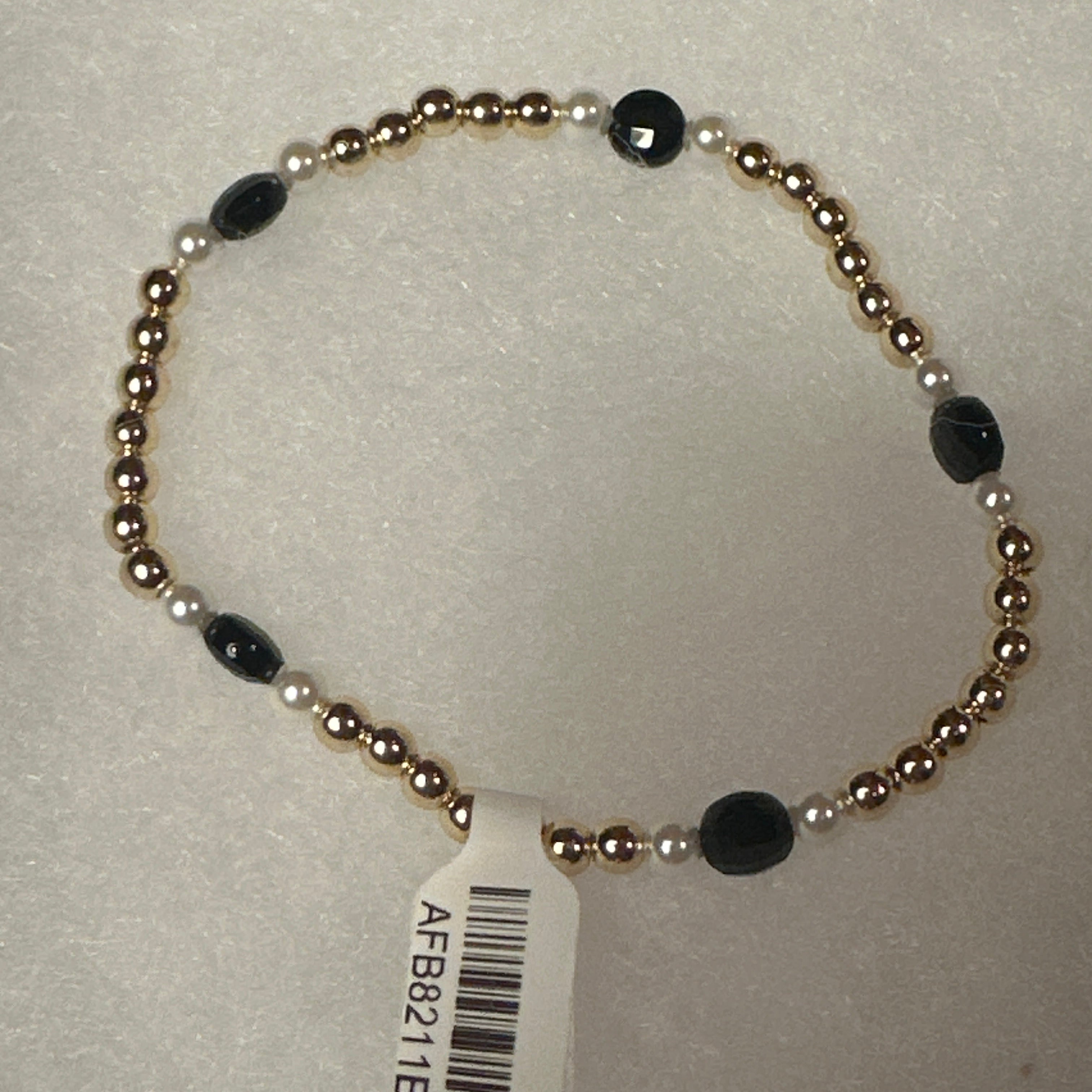 A beaded stretch bracelet with metallic gold-toned round beads, small white pearl-like beads, and faceted black oval beads, shown with a white barcode tag on a light textured background.