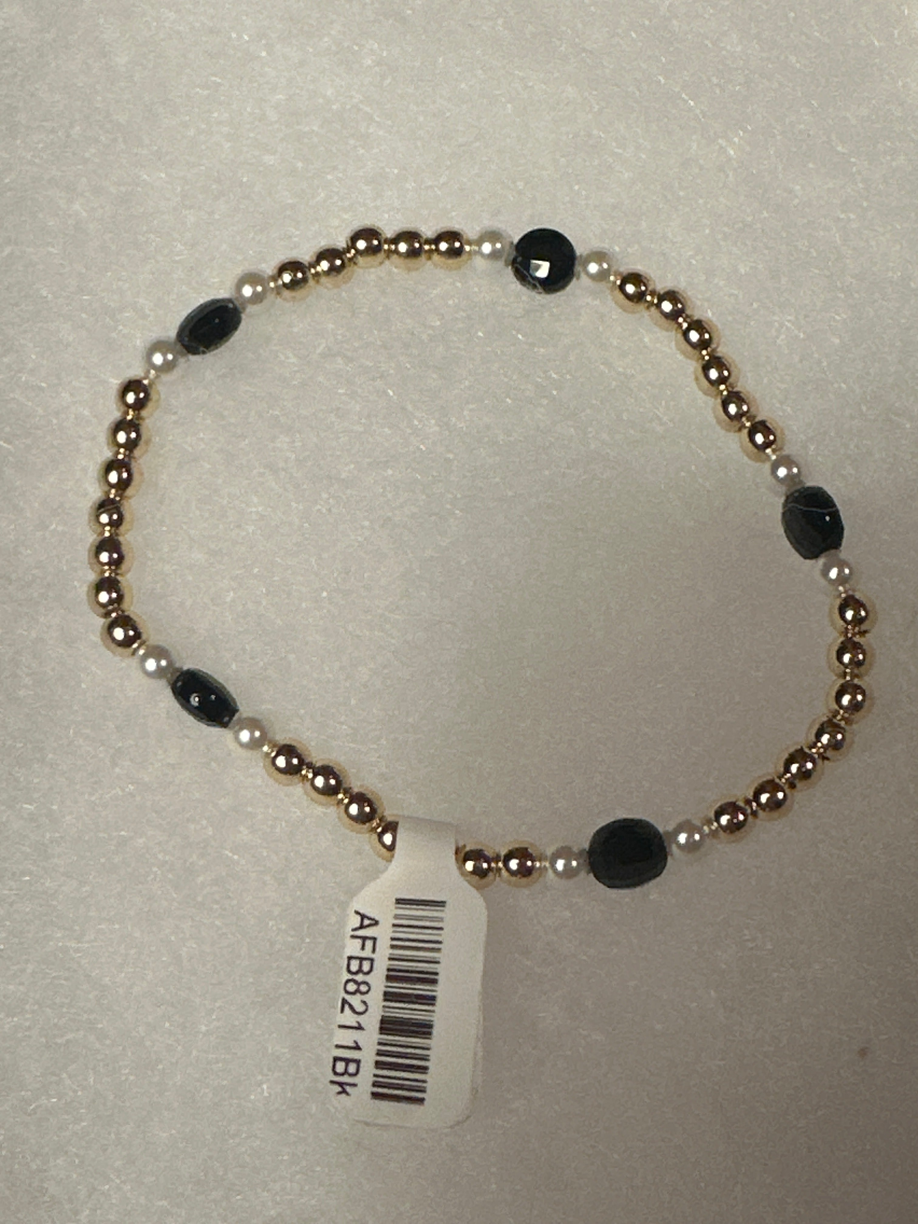 A beaded stretch bracelet with metallic gold-toned round beads, small white pearl-like beads, and faceted black oval beads, shown with a white barcode tag on a light textured background.