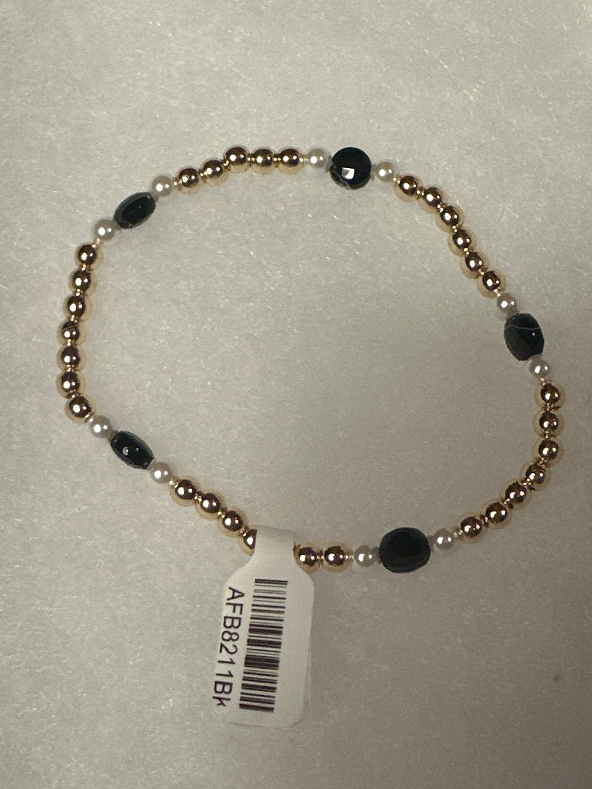 A beaded stretch bracelet with metallic gold-toned round beads, small white pearl-like beads, and faceted black oval beads, shown with a white barcode tag on a light textured background.