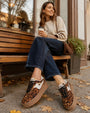 A woman sits on a wooden bench outdoors wearing leopard-print platform sneakers with white side panels, dark blue jeans, and a beige sweater, holding a takeaway coffee cup.