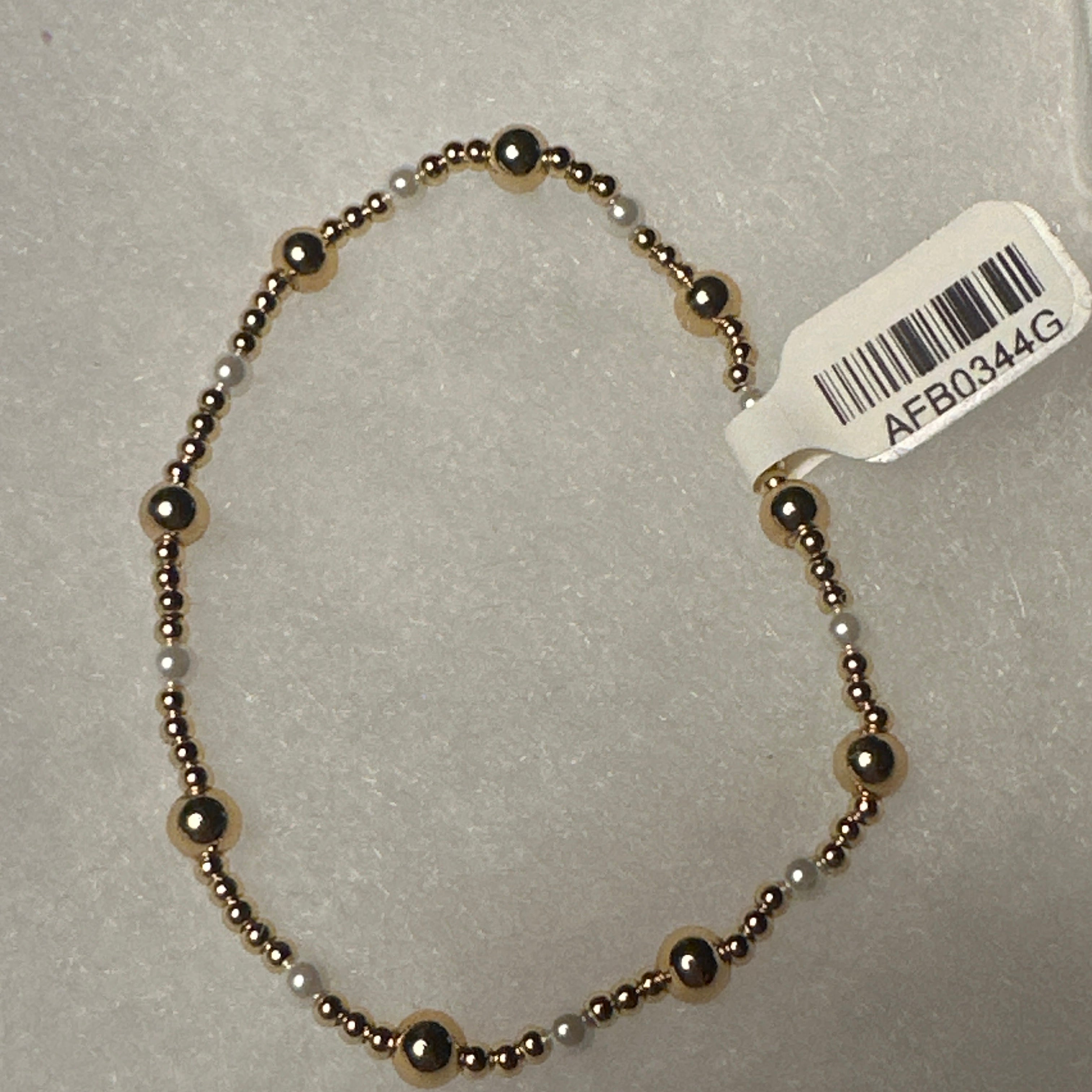 A beaded stretch bracelet with alternating metallic bronze and white beads, shown on a light gray textured background with a white product tag attached.