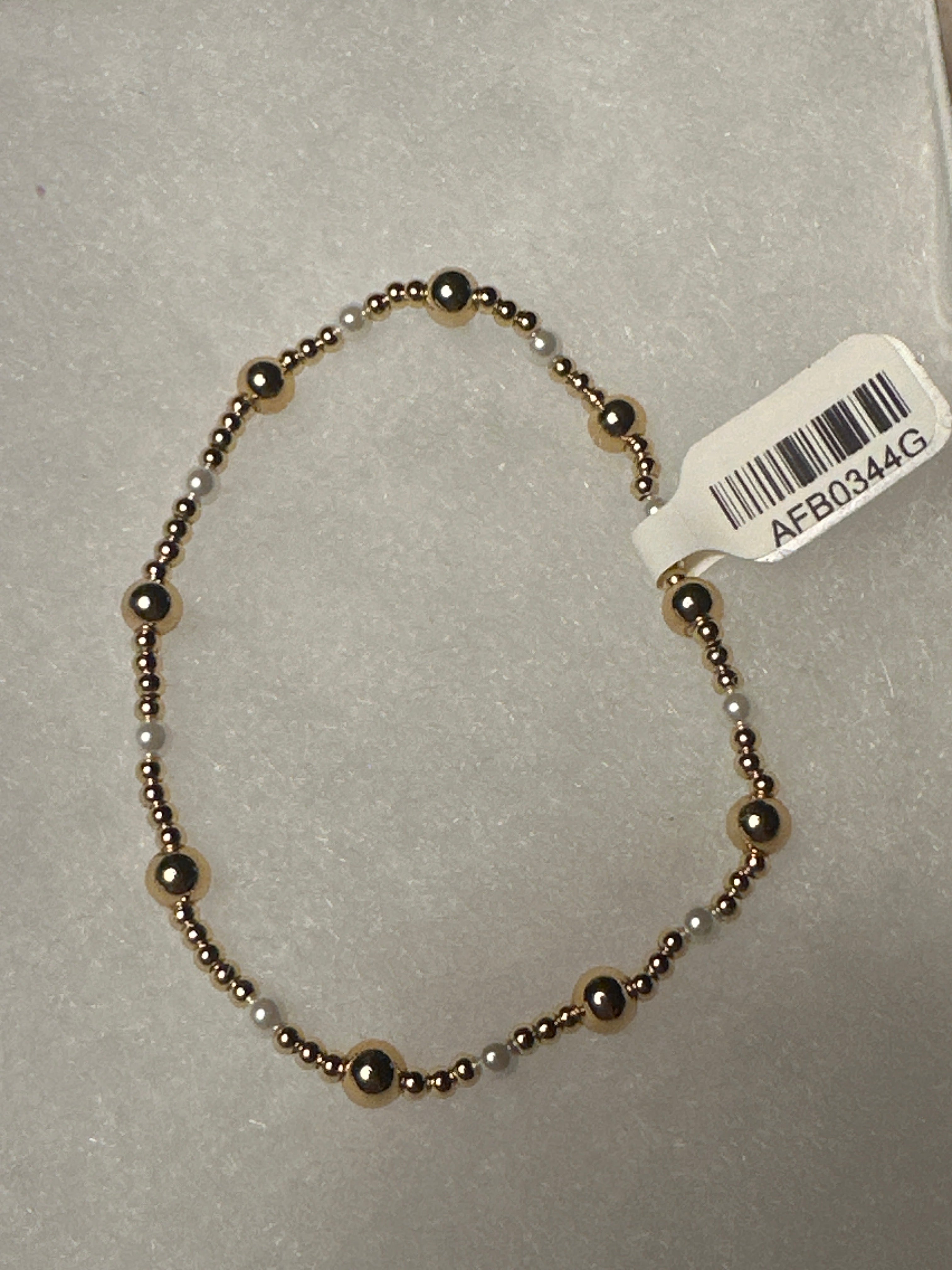 A beaded stretch bracelet with alternating metallic bronze and white beads, shown on a light gray textured background with a white product tag attached.