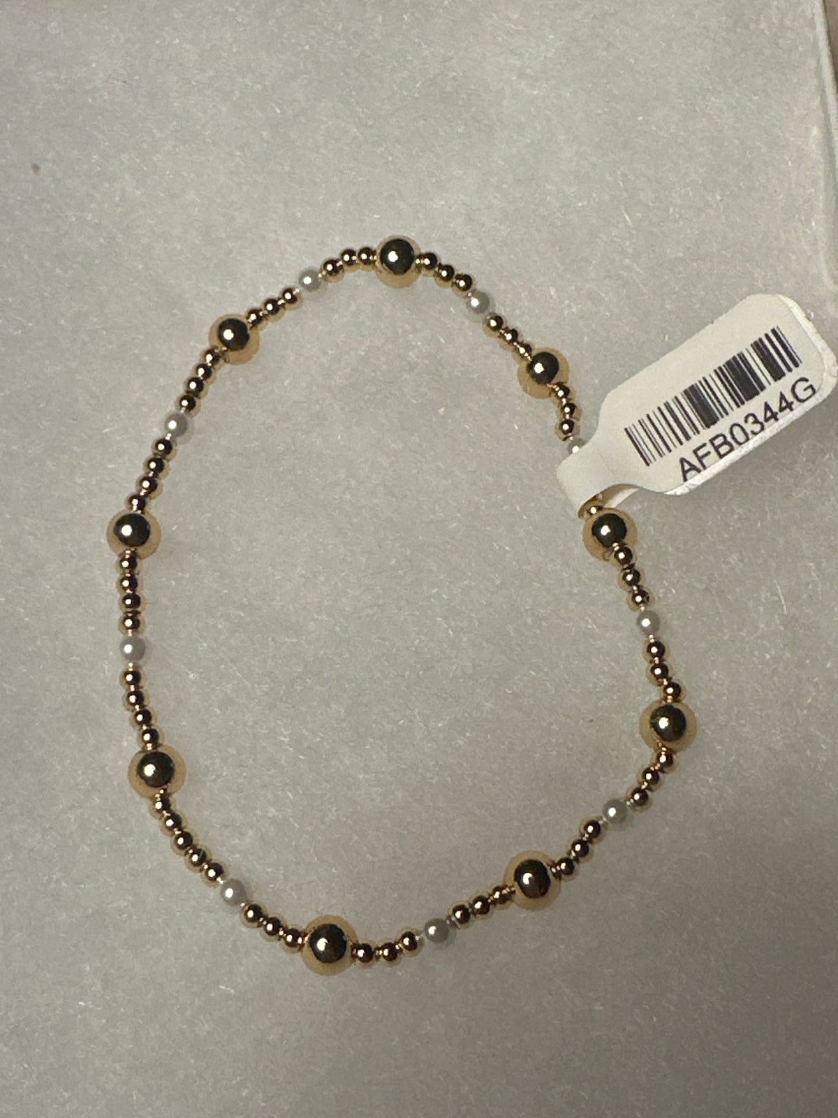 A beaded stretch bracelet with alternating metallic bronze and white beads, shown on a light gray textured background with a white product tag attached.