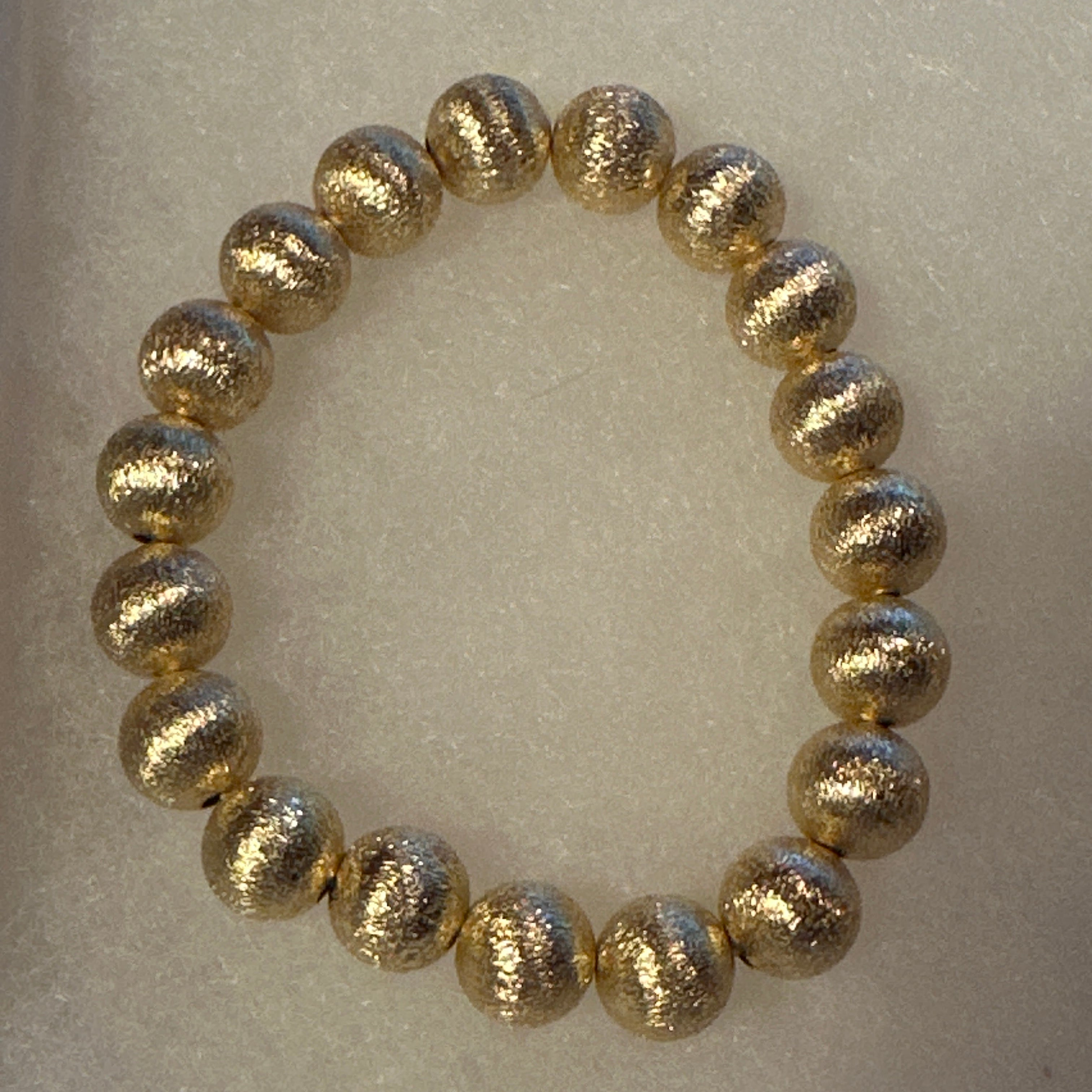 A beaded stretch bracelet made of metallic gold textured round beads, displayed on a light beige textured background.