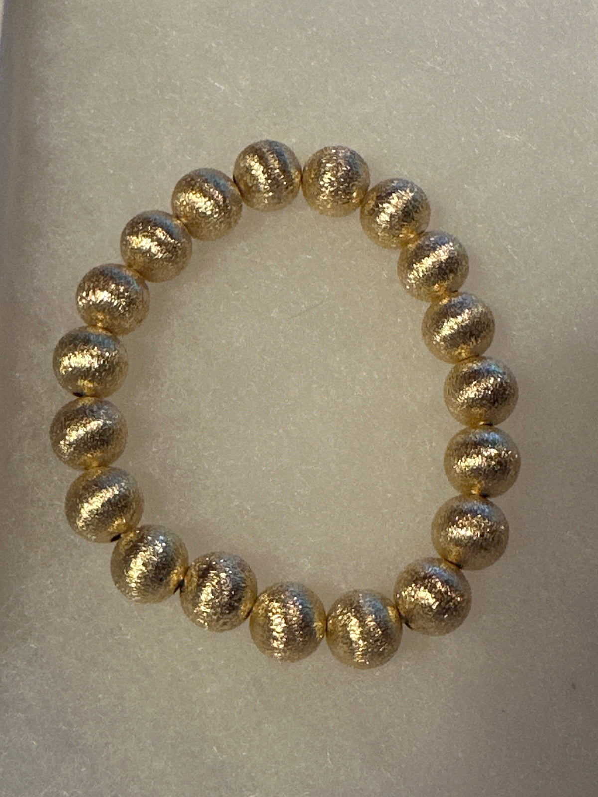 A beaded stretch bracelet made of metallic gold textured round beads, displayed on a light beige textured background.