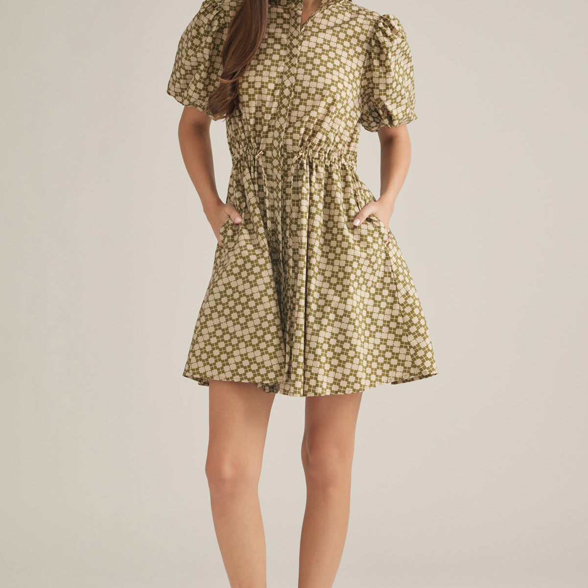 A woman modeling a short puff-sleeve dress with a beige and brown floral pattern, standing against a plain light beige studio background.
