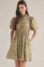 A woman modeling a short puff-sleeve dress with a gathered waist and side pockets, featuring an olive green and beige geometric pattern, against a solid light beige background.