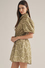 A woman modeling a short puff-sleeve dress with a high mock neck and a beige and olive green geometric pattern, set against a plain light gray studio background.