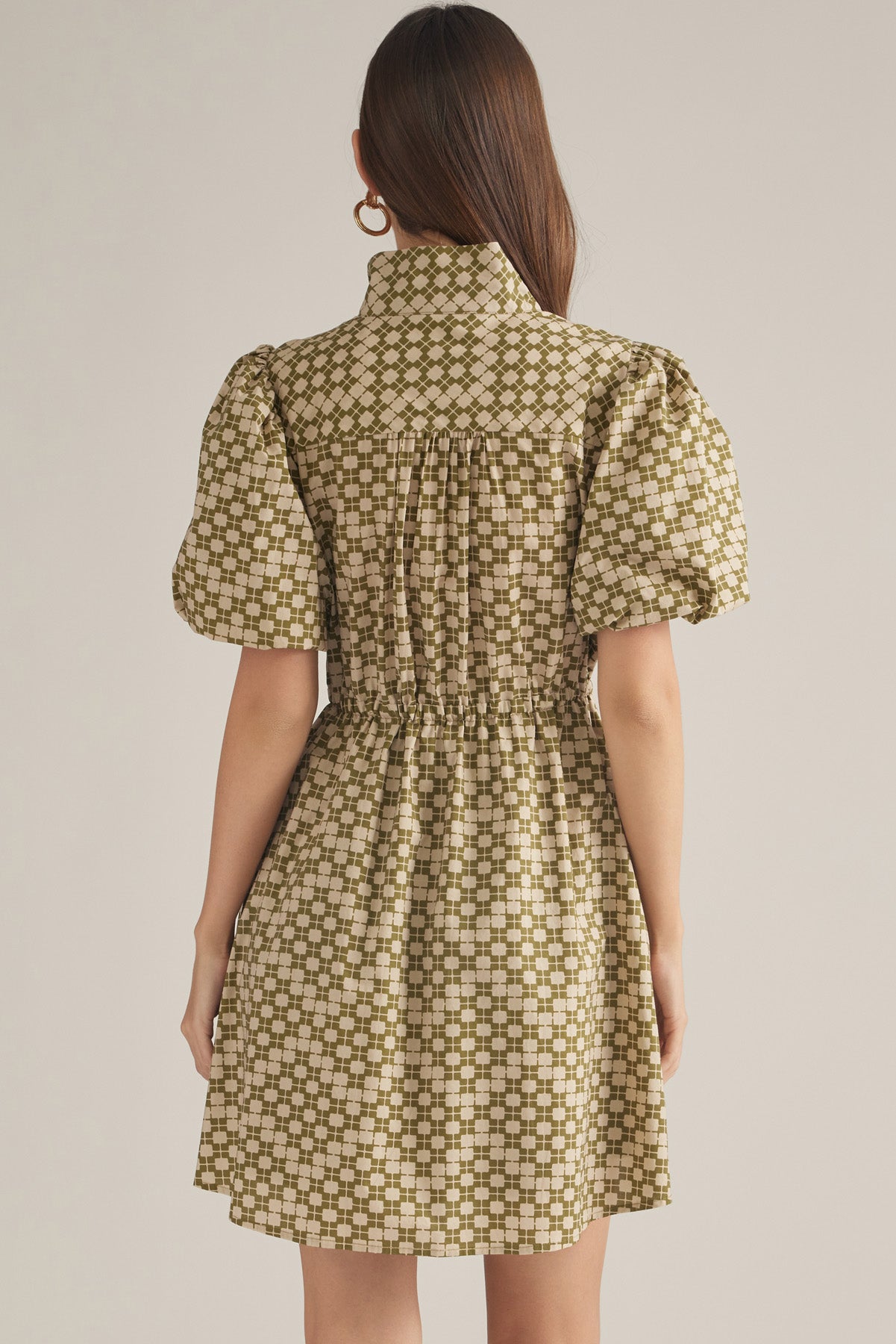 A woman viewed from the back wearing a short puff-sleeve dress with a gathered waist and an allover geometric pattern in olive green and cream on a light beige background.