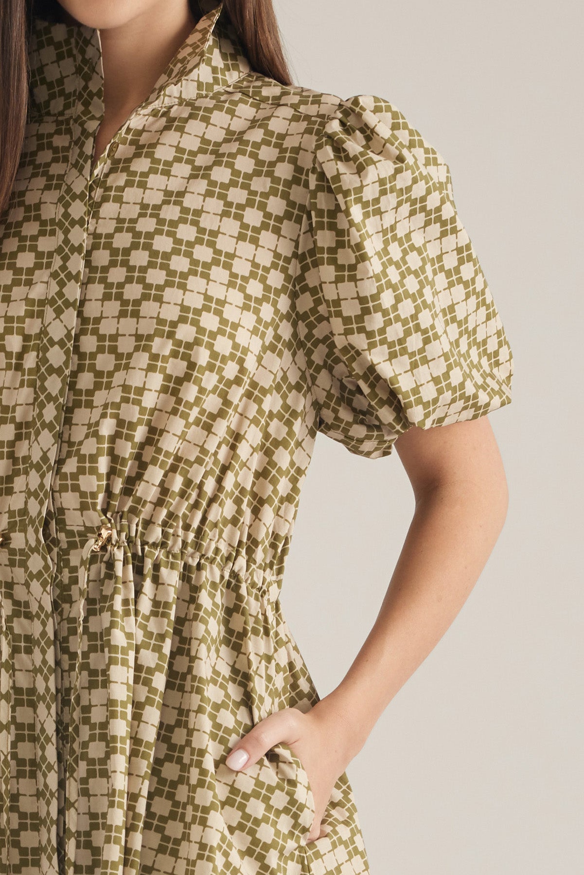 A close-up of a model wearing a short-sleeve dress with puff sleeves and a gathered waist, featuring an olive green and cream checkered pattern against a solid light gray background.