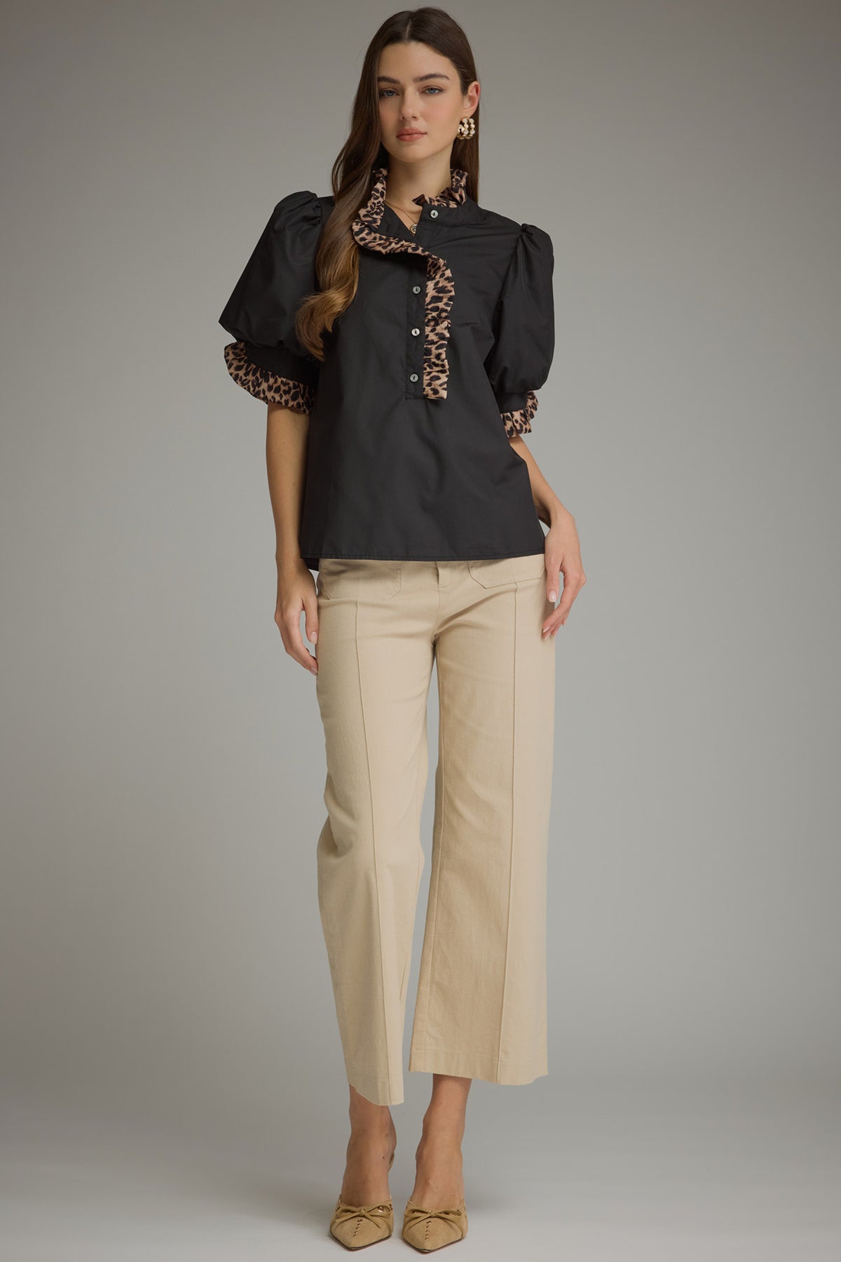 A woman modeling a black puff-sleeve blouse with leopard-print trim and buttons, paired with beige cropped trousers and beige pointed flats, against a plain gray studio background.