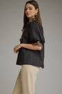 A woman modeling a black short-sleeve blouse with leopard-print ruffle trim at the collar and cuffs, paired with beige trousers, against a plain gray studio background.