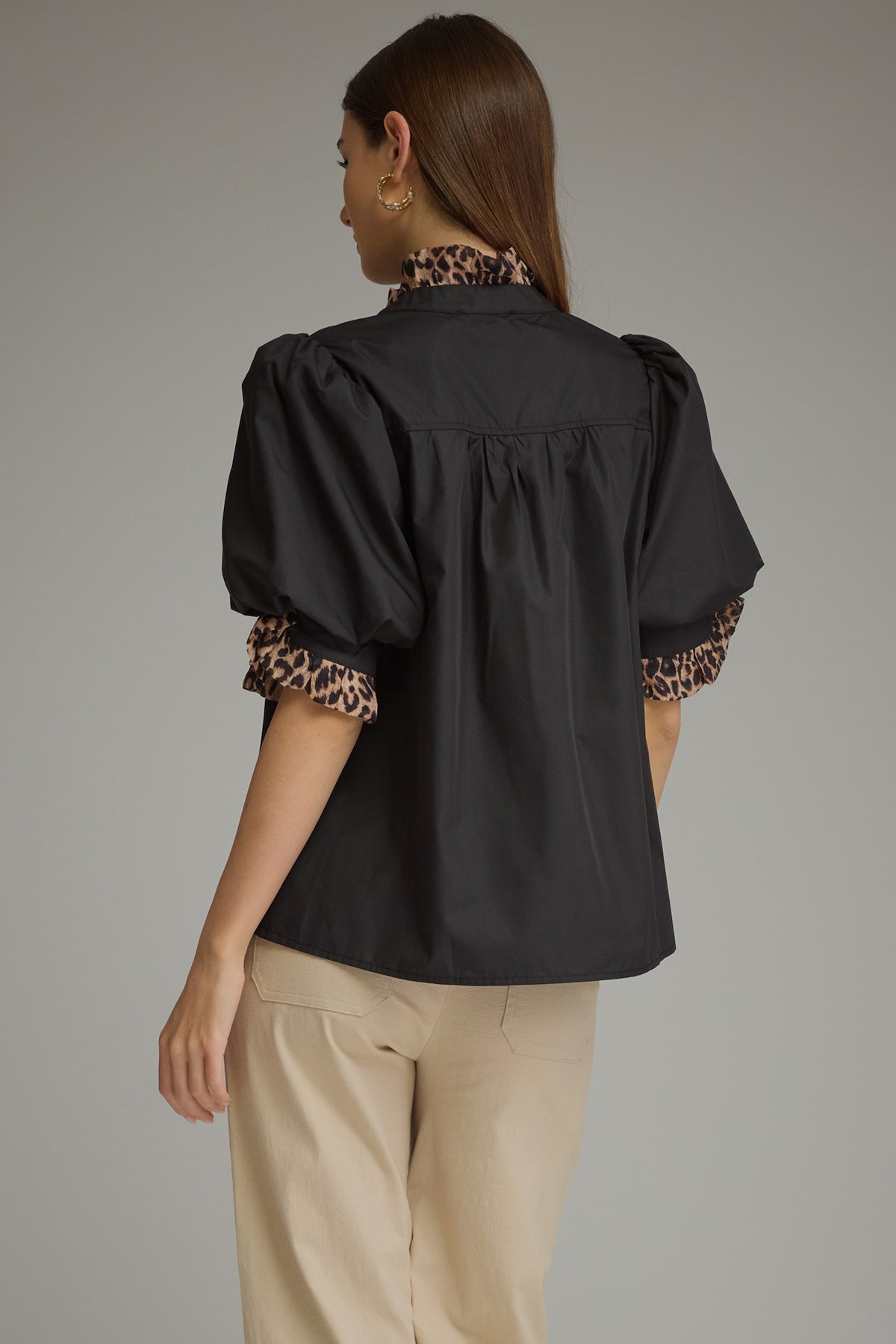 A woman shown from the back wearing a black puff-sleeve blouse with leopard-print trim at the collar and sleeve cuffs, paired with beige trousers, against a plain gray studio background.