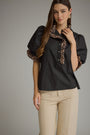 A woman modeling a black short-sleeve blouse with leopard-print trim on the collar and sleeve cuffs, paired with beige pants, against a solid gray studio background.