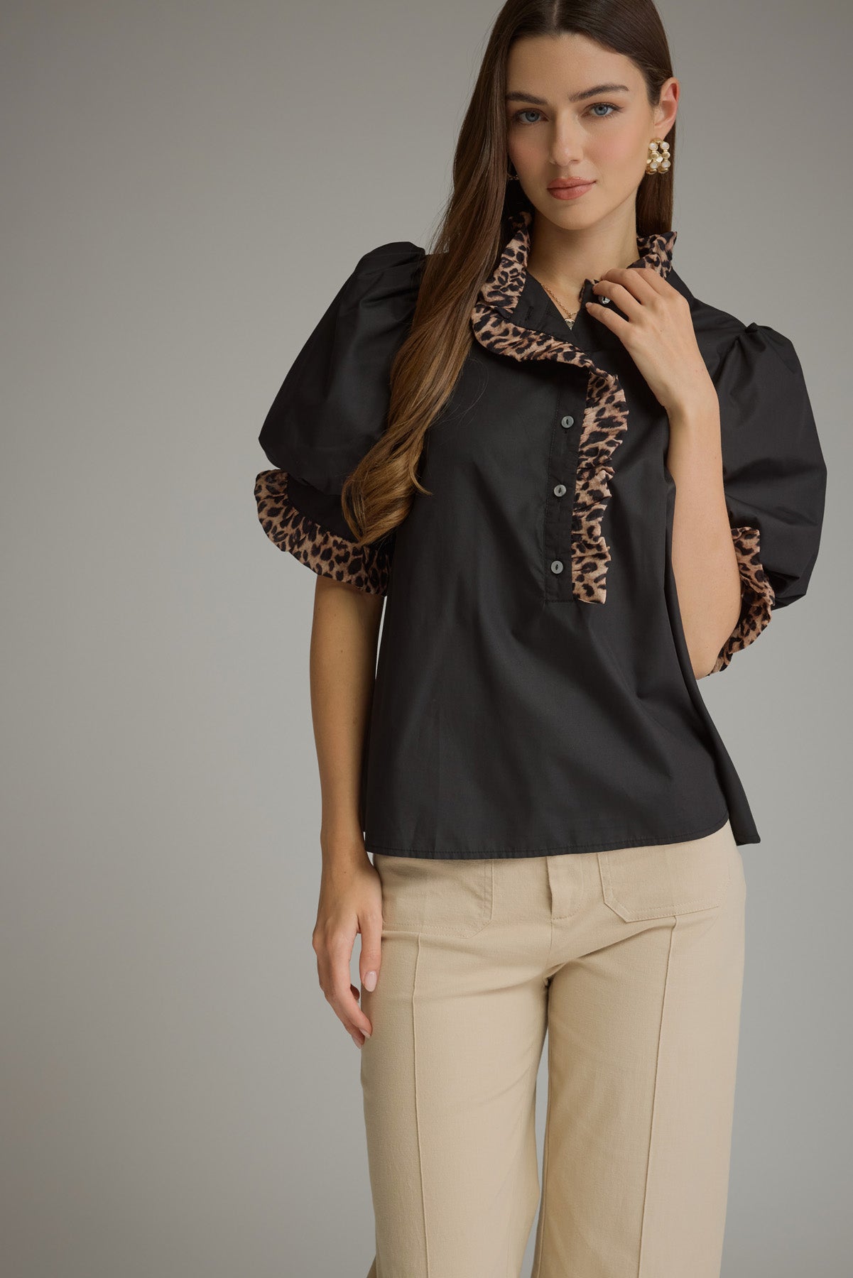 A woman modeling a black short-sleeve blouse with leopard-print trim on the collar and sleeve cuffs, paired with beige pants, against a solid gray studio background.