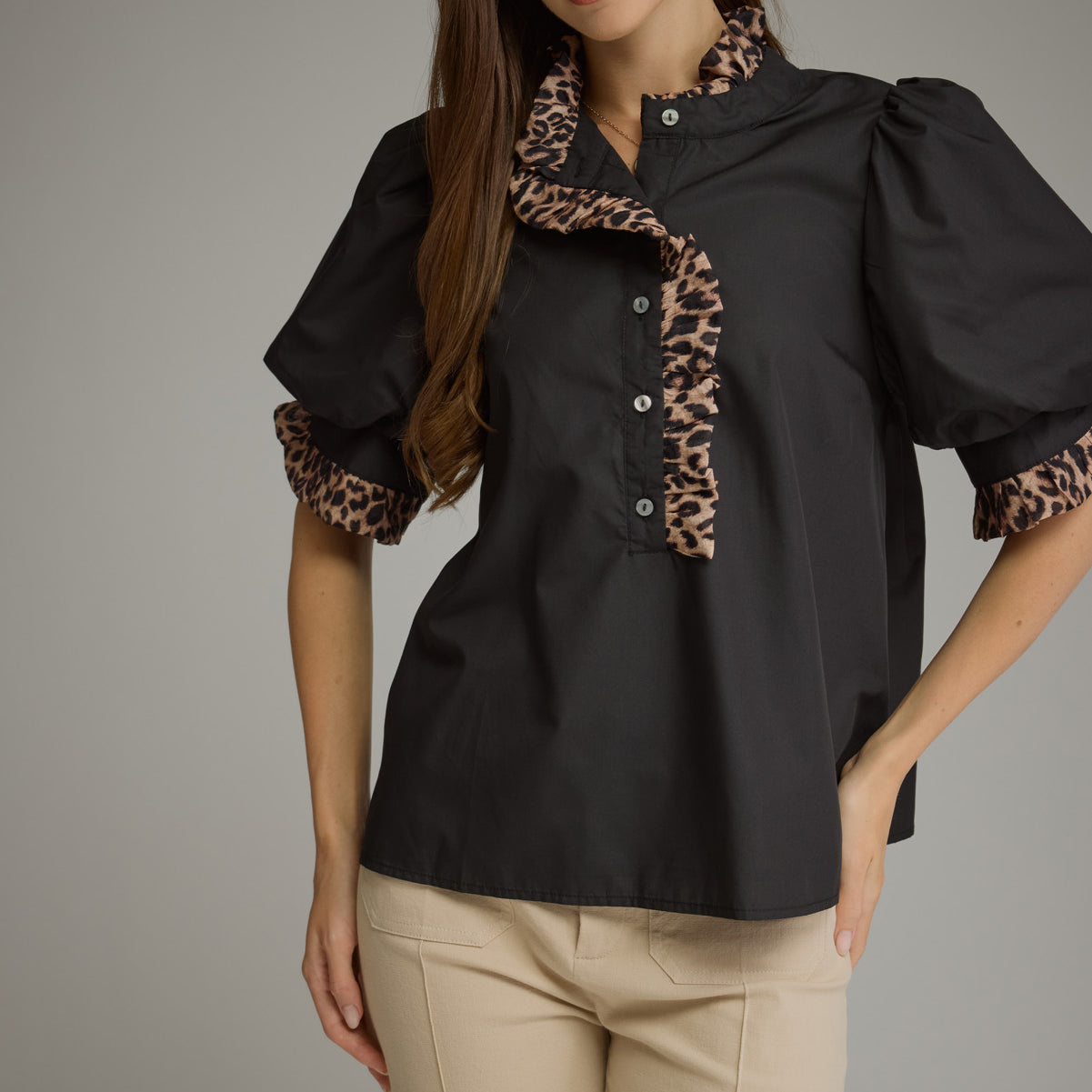 A woman modeling a black short-sleeve blouse with leopard-print ruffle trim on the collar and sleeve cuffs, paired with beige pants, against a plain gray studio background.
