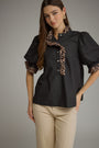 A woman modeling a black short-sleeve blouse with leopard-print ruffle trim on the collar and sleeve cuffs, paired with beige pants, against a plain gray studio background.
