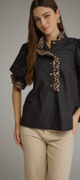 A woman modeling a black short-sleeve blouse with leopard-print ruffle trim on the collar and sleeve cuffs, paired with beige pants, against a plain gray studio background.