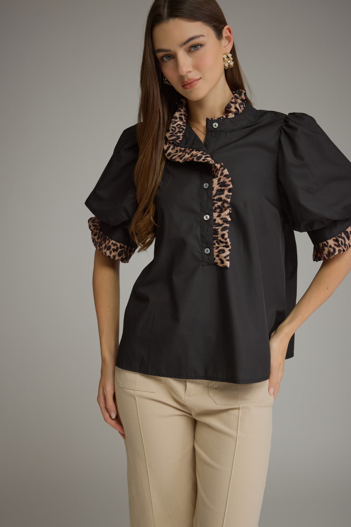 A woman modeling a black short-sleeve blouse with leopard-print ruffle trim on the collar and sleeve cuffs, paired with beige pants, against a plain gray studio background.