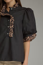 A close-up of a woman wearing a black blouse with leopard-print ruffled trim on the collar, placket, and sleeve cuffs, against a plain gray studio background.