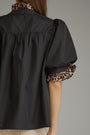 A close-up back view of a model wearing a black short-sleeve blouse with leopard-print ruffled trim on the collar and cuffs, paired with beige trousers, against a solid gray background.