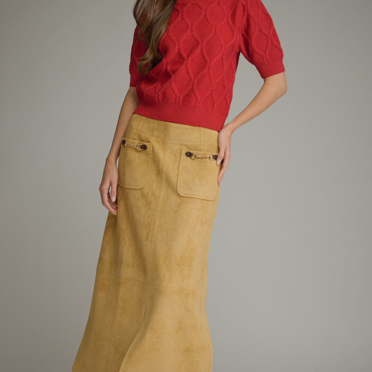 A woman modeling a red short-sleeve textured knit top paired with a mustard yellow suede skirt with front patch pockets, set against a plain gray studio background.