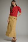 A woman modeling a red short-sleeve textured knit top paired with a mustard yellow suede skirt with front patch pockets, set against a plain gray studio background.