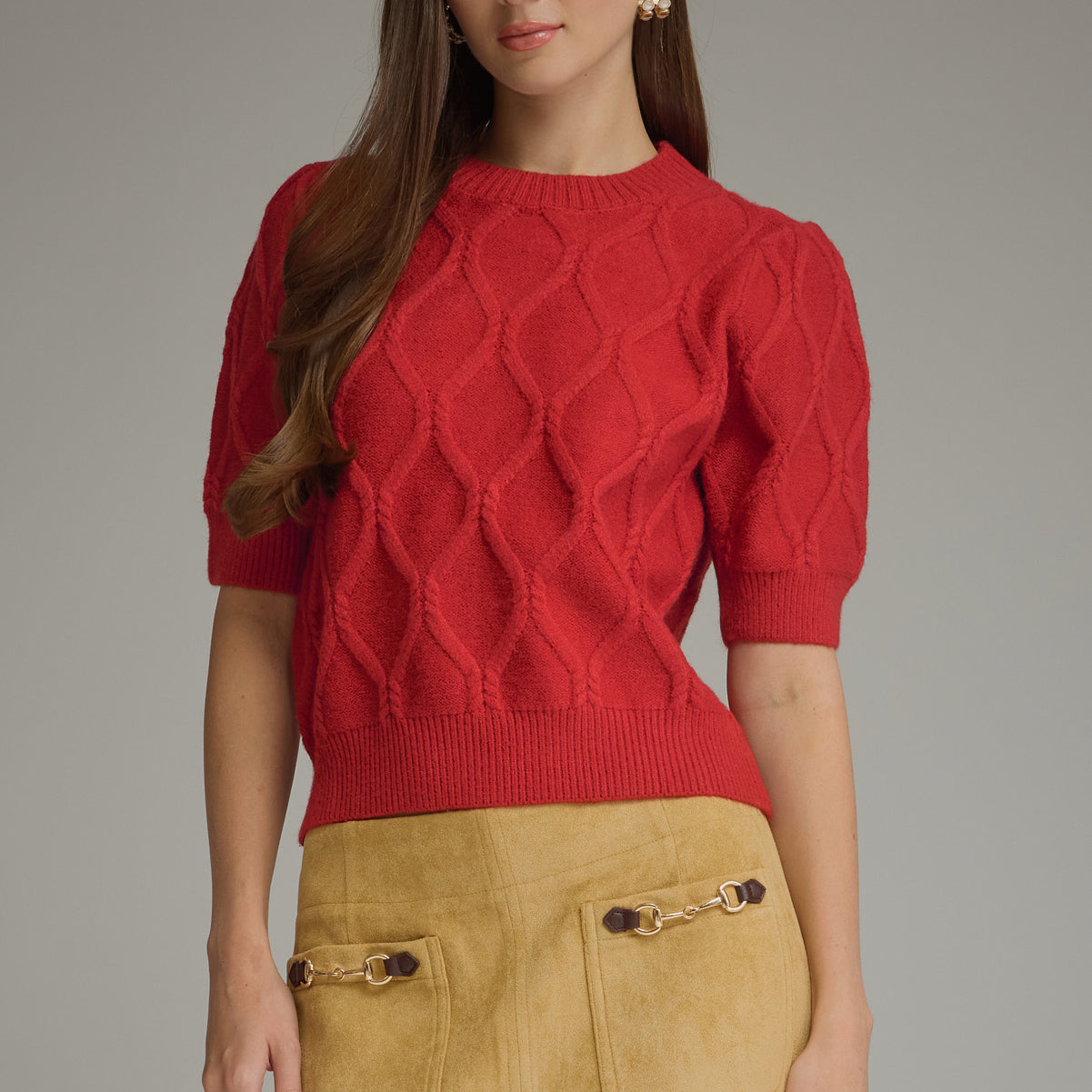 A woman modeling a red short-sleeve knit sweater with a textured diamond pattern, paired with a tan suede skirt, against a solid gray studio background.