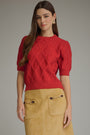 A woman modeling a red short-sleeve knit sweater with a textured diamond pattern, paired with a tan suede skirt, against a solid gray studio background.