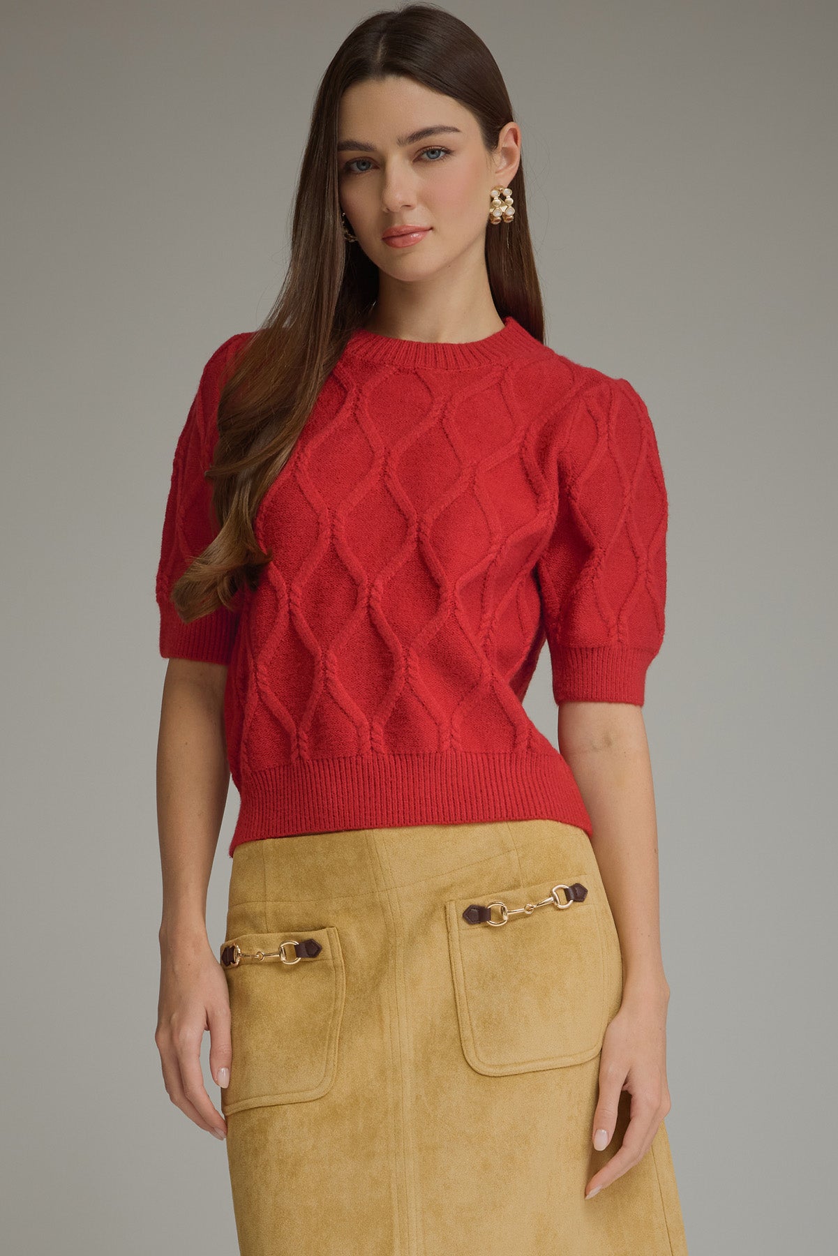 A woman modeling a red short-sleeve knit sweater with a textured diamond pattern, paired with a tan suede skirt, against a solid gray studio background.
