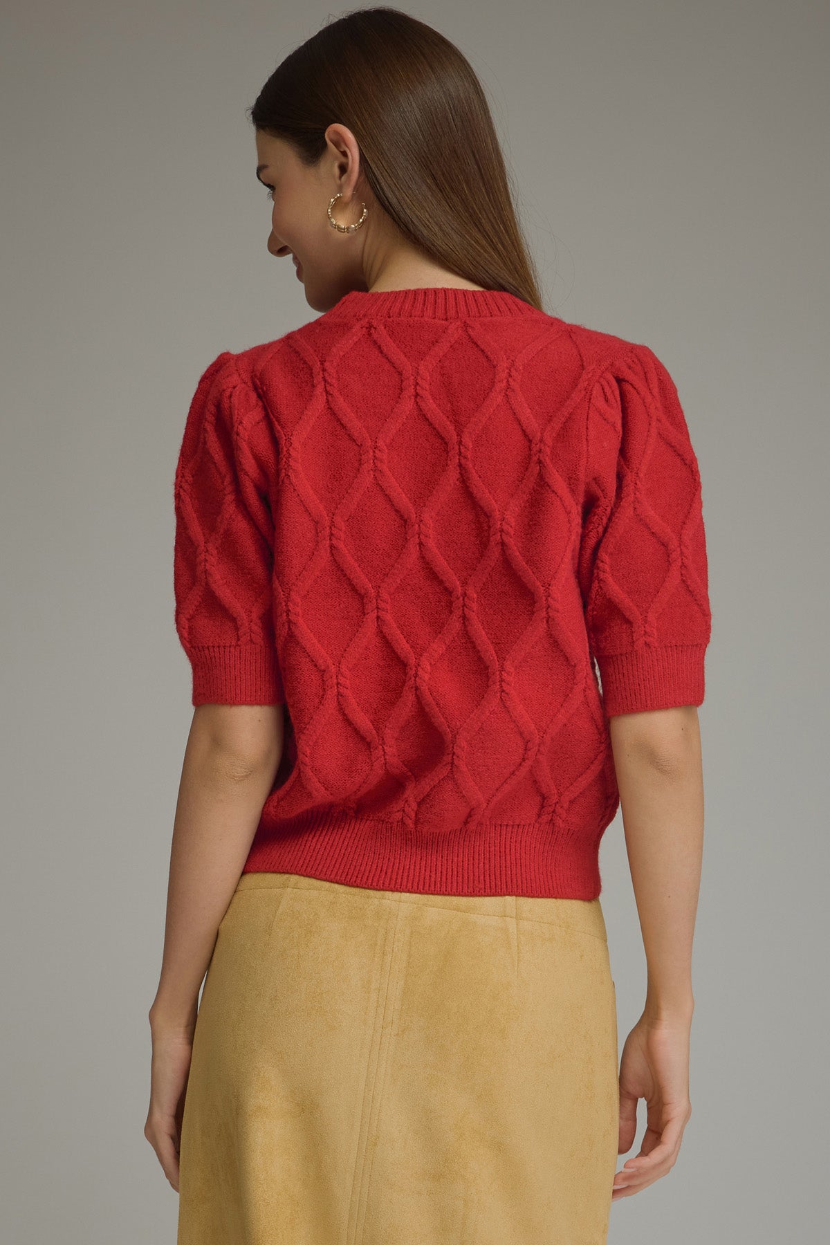 A woman shown from the back wearing a bright red short-sleeve knit top with a textured diamond pattern and a beige skirt, against a plain gray studio background.