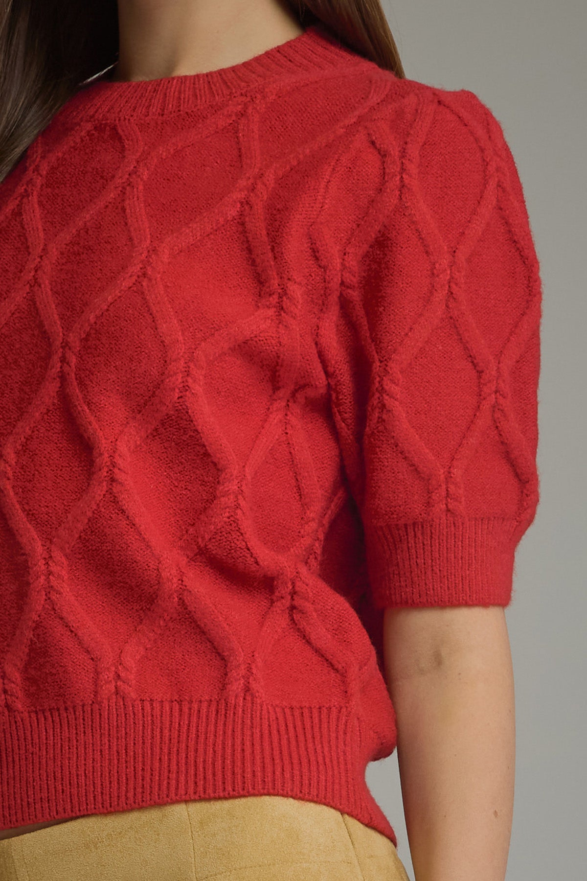A close-up of a person wearing a bright red short-sleeve knit sweater with a textured cable pattern and ribbed hem, paired with a beige skirt.