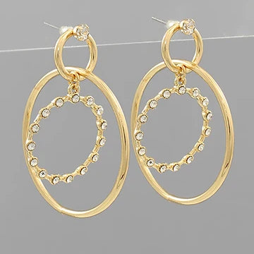 A pair of gold-toned drop earrings with large oval hoops and a smaller inner oval outlined with small clear rhinestones, shown against a plain gray background.