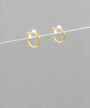 A pair of small gold hoop earrings with white gemstone accents, suspended on a thin white wire against a solid gray background.