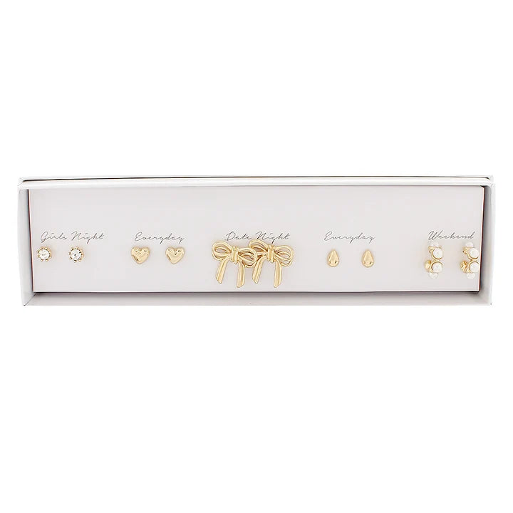 A set of five gold-toned stud earrings displayed in a white rectangular presentation box against a white background. The earrings include different designs such as a heart, a bow, teardrop, and floral motifs, with some labeled with handwritten text like 'Girls Night' and 'Everyday'.