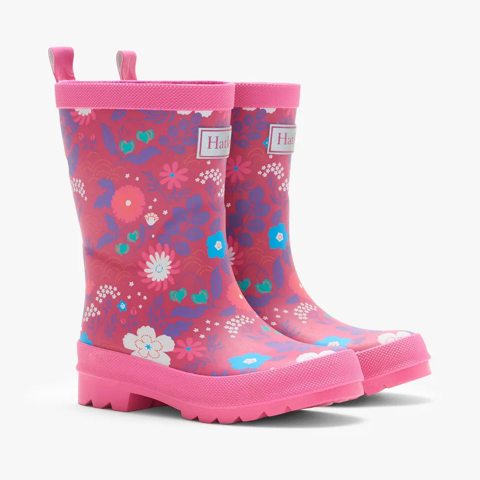 A pair of children's pink rubber rain boots with a colorful floral and abstract pattern, featuring pull tabs and a thick lug sole, shown on a light gray background.