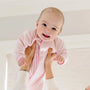 A baby wearing a light pink zip-up sleeper is being held up by an adult's hands in a bright, clean indoor setting.
