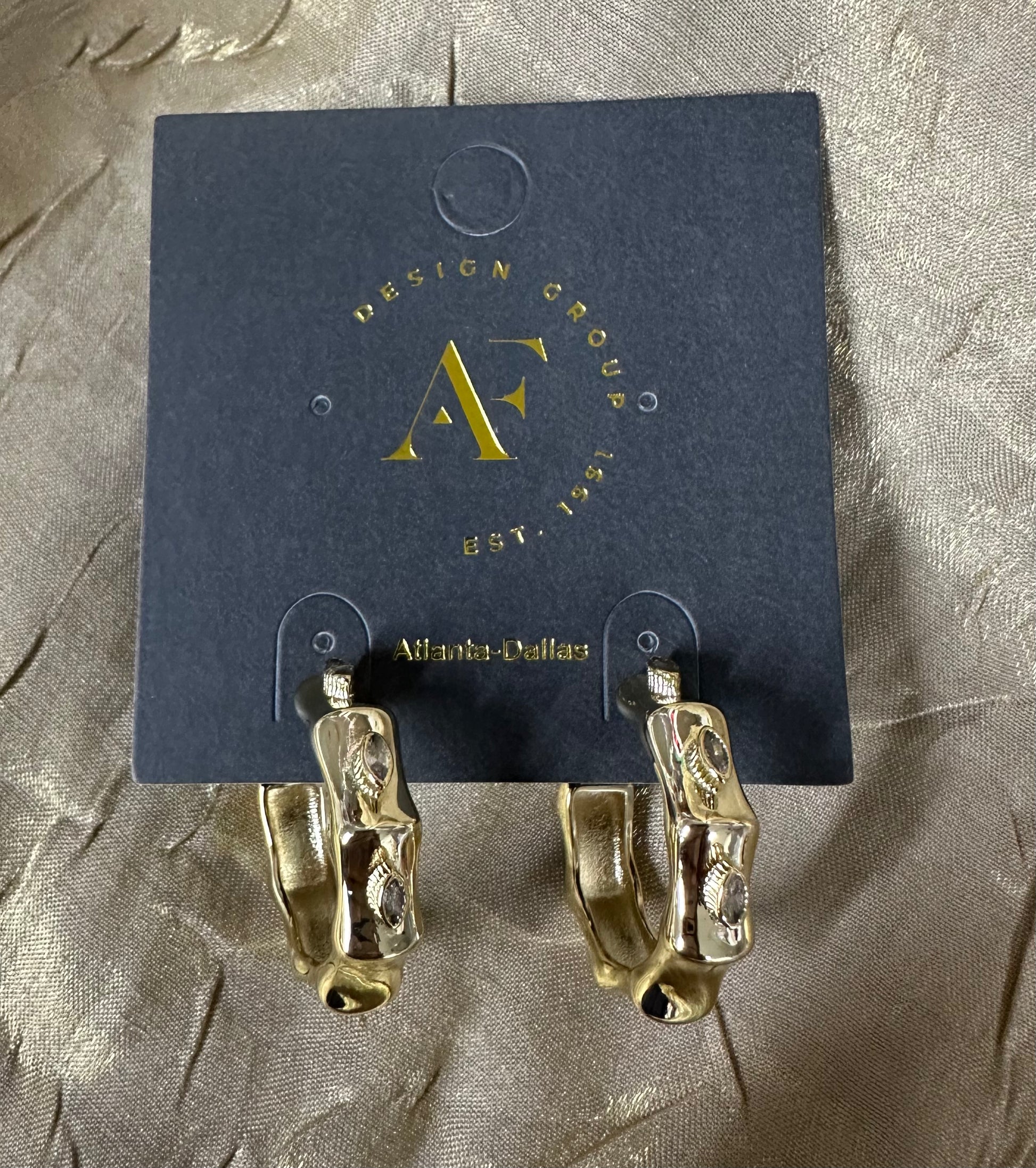 A pair of gold hoop earrings with a textured, organic pattern, displayed on a dark blue card from Design Group Atlanta. The earrings are photographed on a textured, shimmering taupe fabric background.