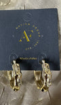 A pair of gold hoop earrings with a textured, organic pattern, displayed on a dark blue card from Design Group Atlanta. The earrings are photographed on a textured, shimmering taupe fabric background.