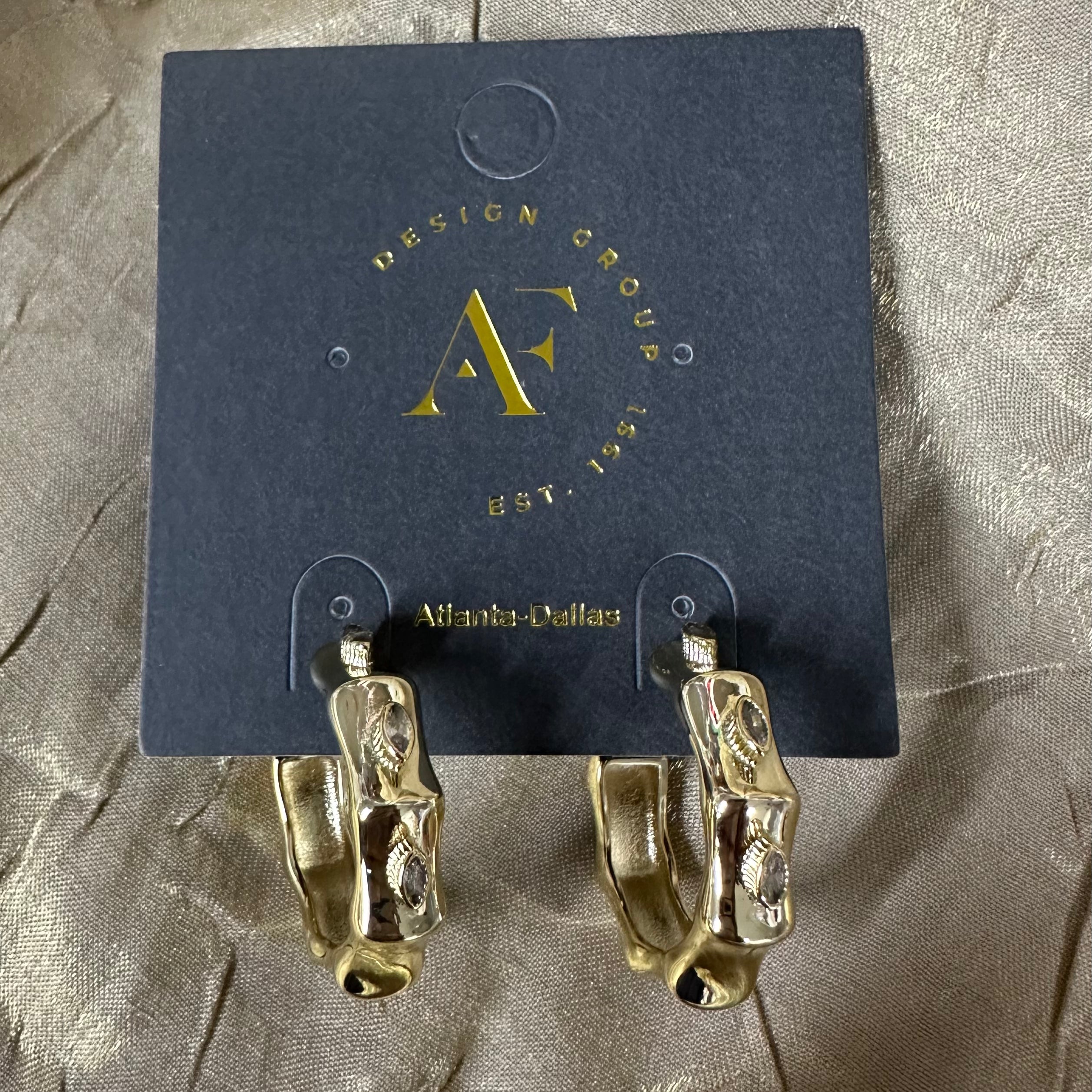 A pair of gold hoop earrings with a textured, organic pattern, displayed on a dark blue card from Design Group Atlanta. The earrings are photographed on a textured, shimmering taupe fabric background.